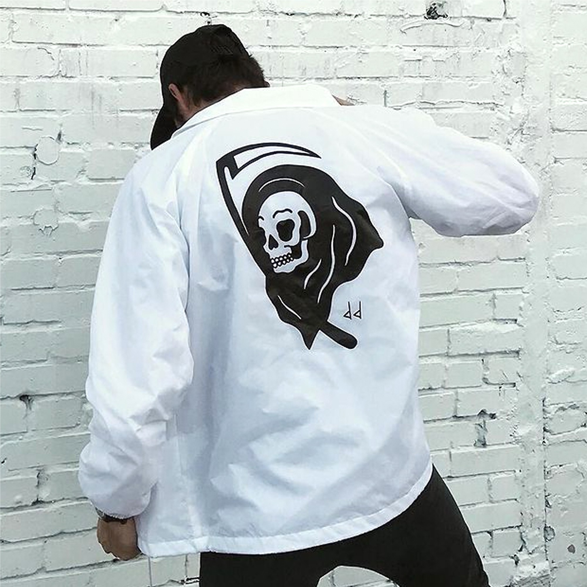 Person wearing a white jacket with a skull and hood design, standing against a white brick wall.