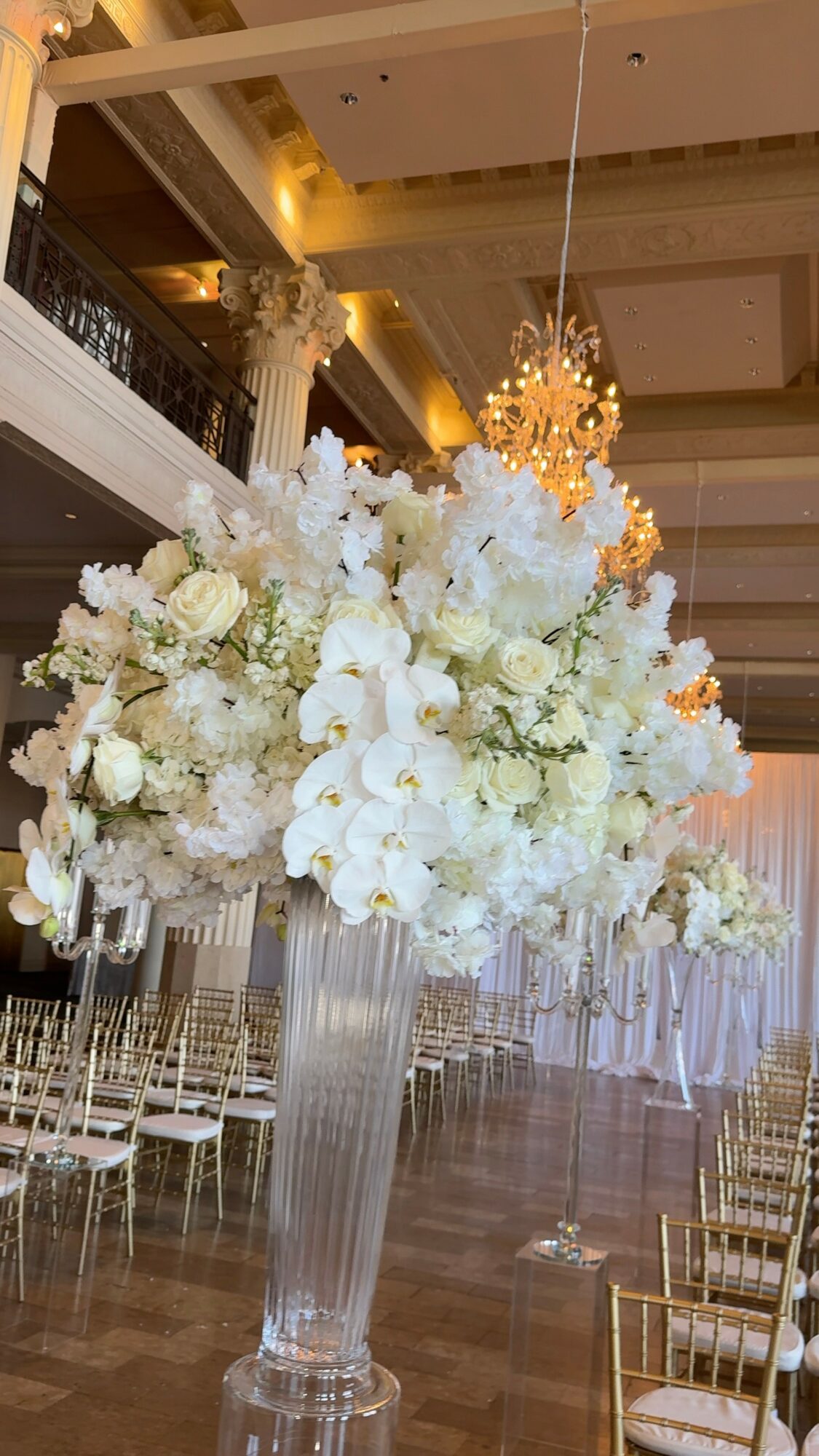 Large floral arrangement with white flowers in a tall glass vase, hanging from the ceiling in an elegant venue.