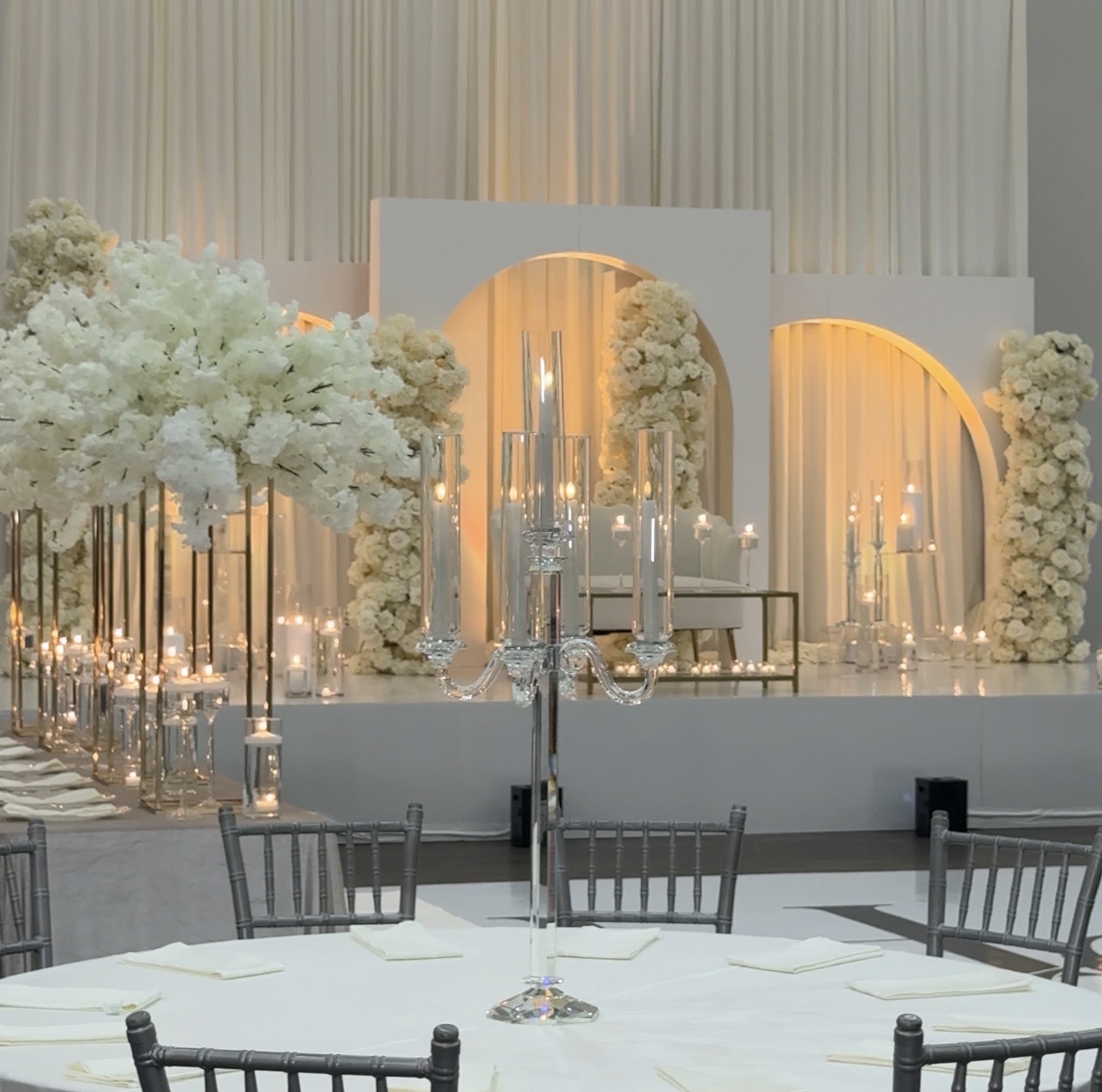 Elegant event setup with floral arrangements, candles, and a decorated stage backdrop, featuring white and gold accents.