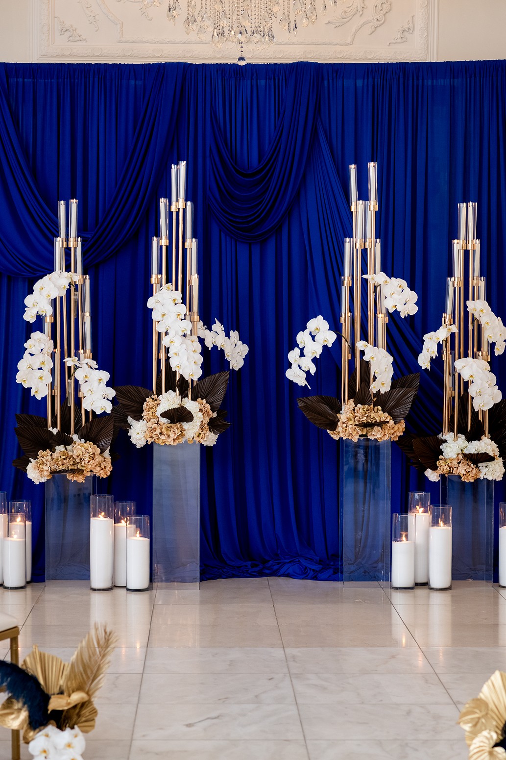 Elegant display with tall white candles, white flowers, and gold accents against a blue curtain backdrop.