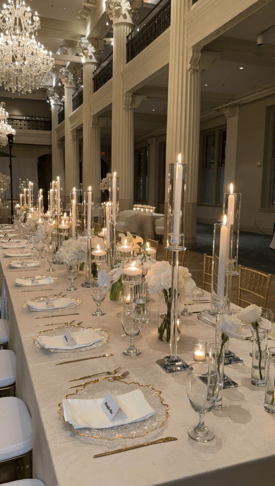 Elegant banquet table with tall candles, floral arrangements, and place settings in a grand hall with chandeliers.