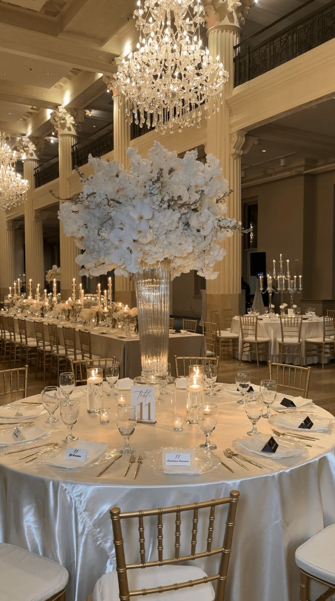 Round table with white tablecloth, tall floral centerpiece, and elegant place settings in a grand ballroom.