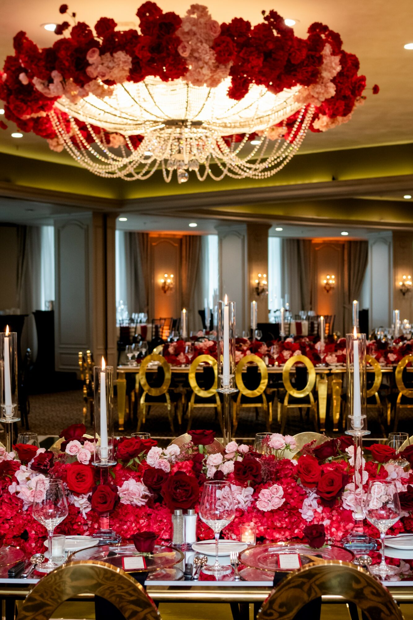 Elegant banquet hall with a large chandelier, decorated with red and pink flowers, and set tables with gold chairs.