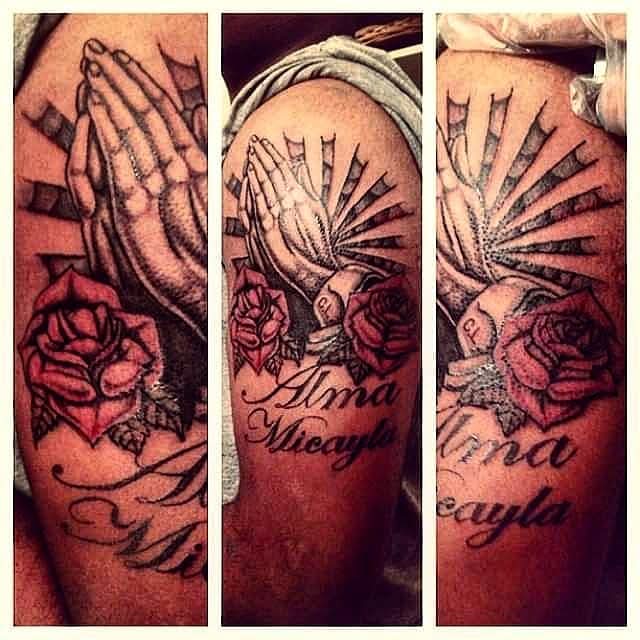 Tattoo of praying hands, roses, and a shell with text, on a person's arm, in black and gray ink.