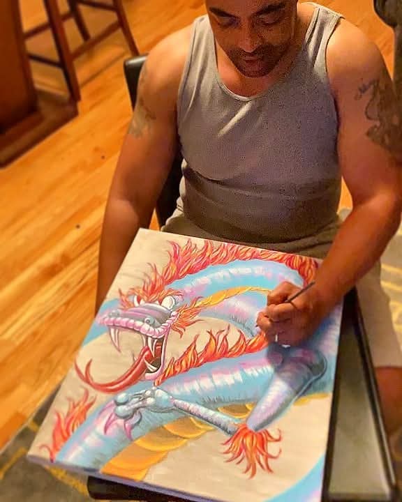 Man in gray tank top sitting at table, showing colorful dragon artwork with flames and clouds, on a wooden floor.