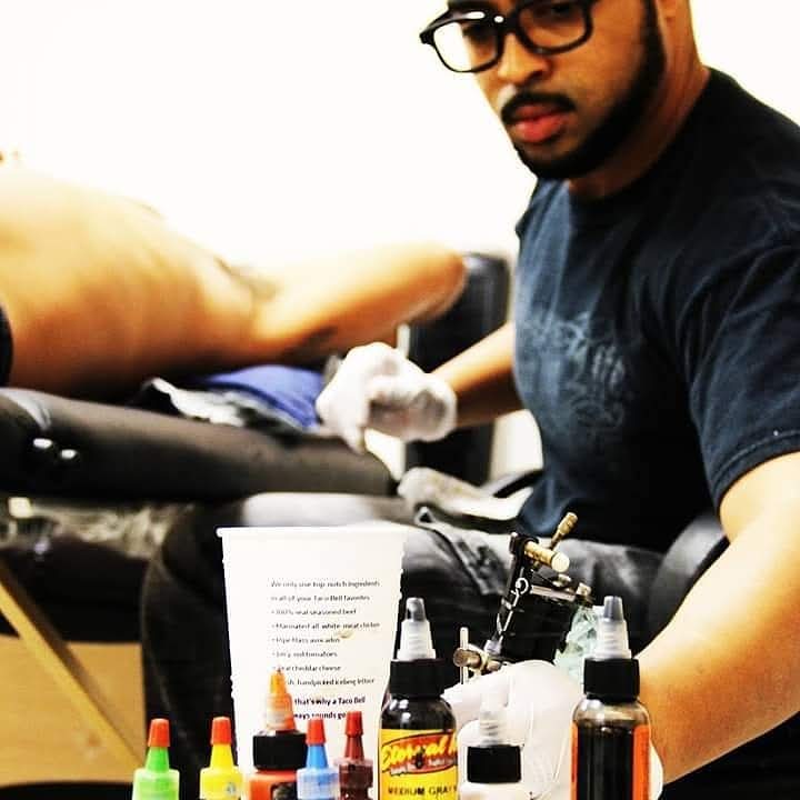 Tattoo artist wearing glasses working on a client's arm with tattoo supplies nearby, in a studio setting.