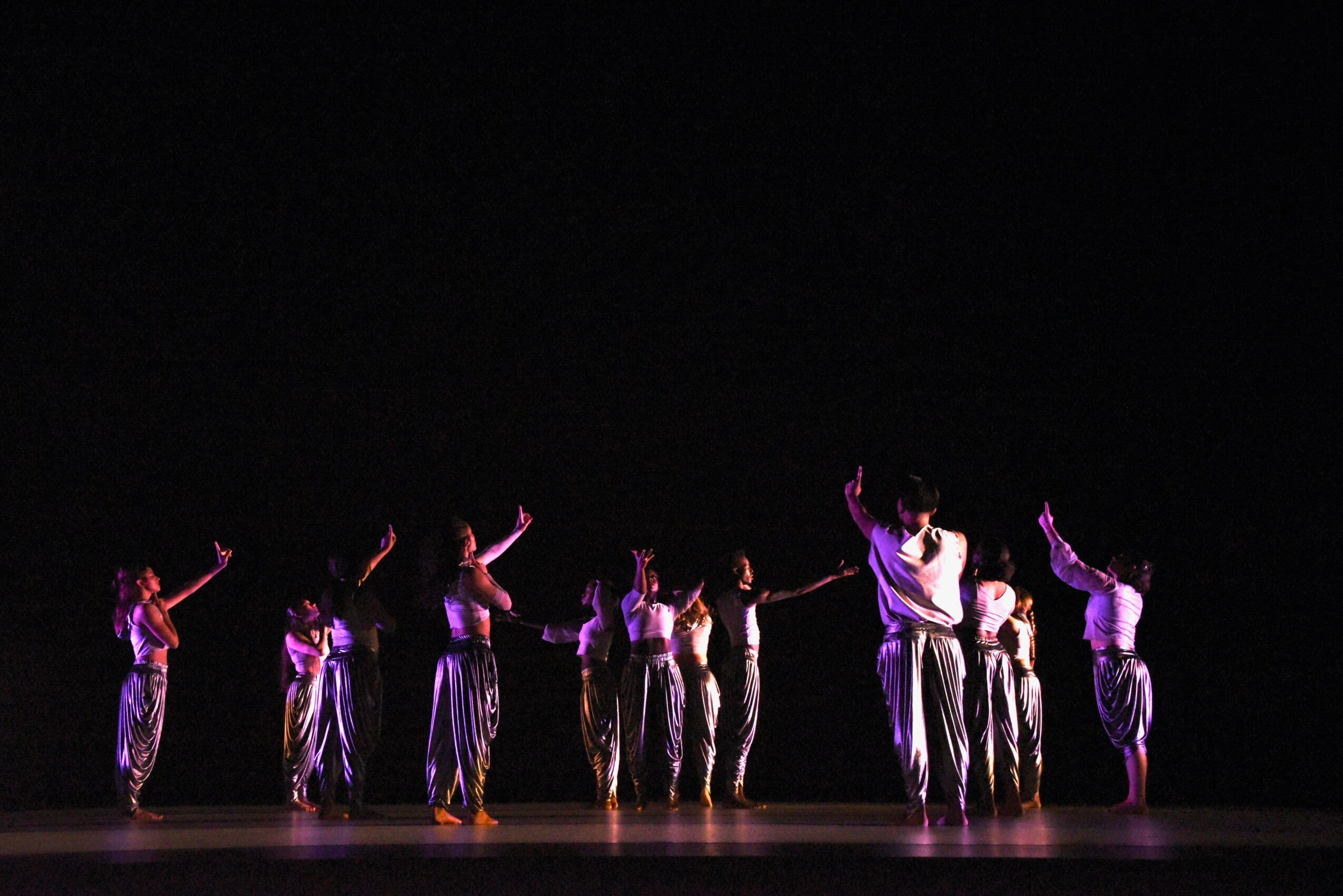 Group of dancers on stage with arms raised, wearing shiny pants, against a dark background.