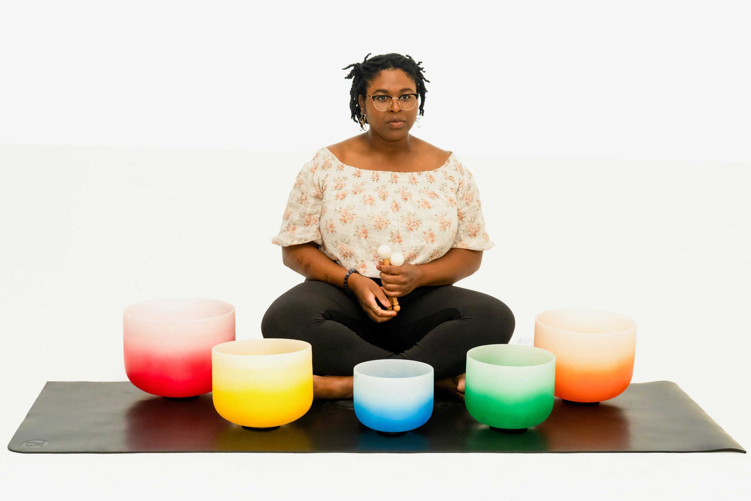 Person sitting cross-legged on a mat with five colorful singing bowls in front, against a plain white background.