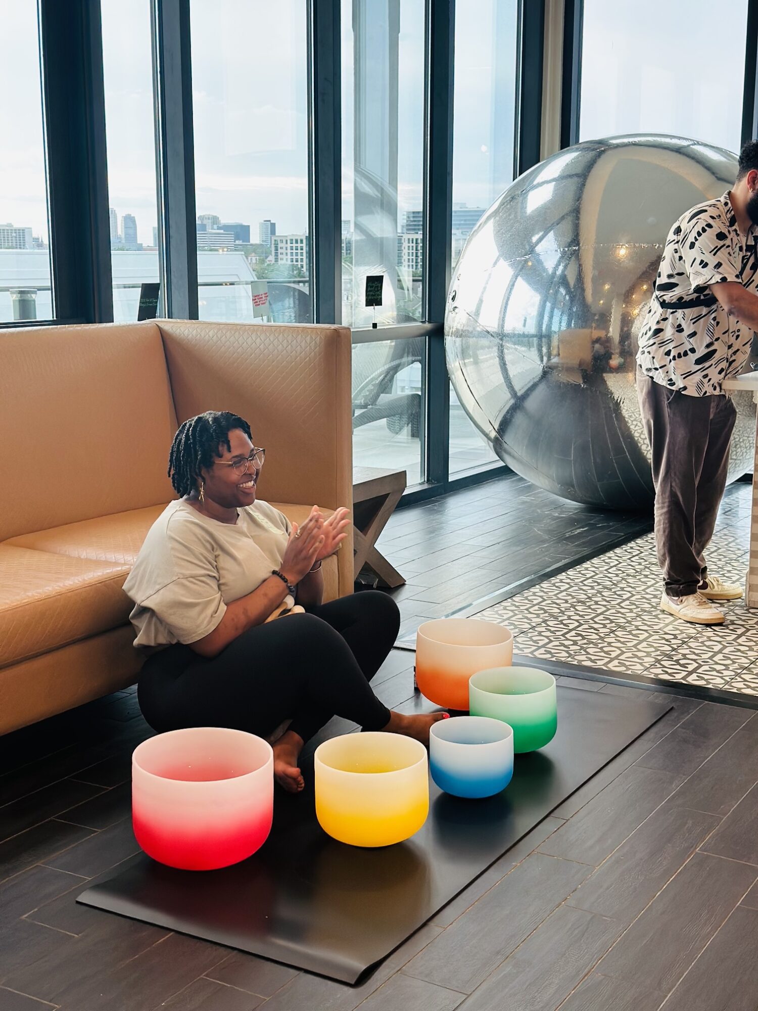 Person sitting on floor with colorful illuminated bowls nearby, in a modern indoor space with large windows and a reflective sphere.