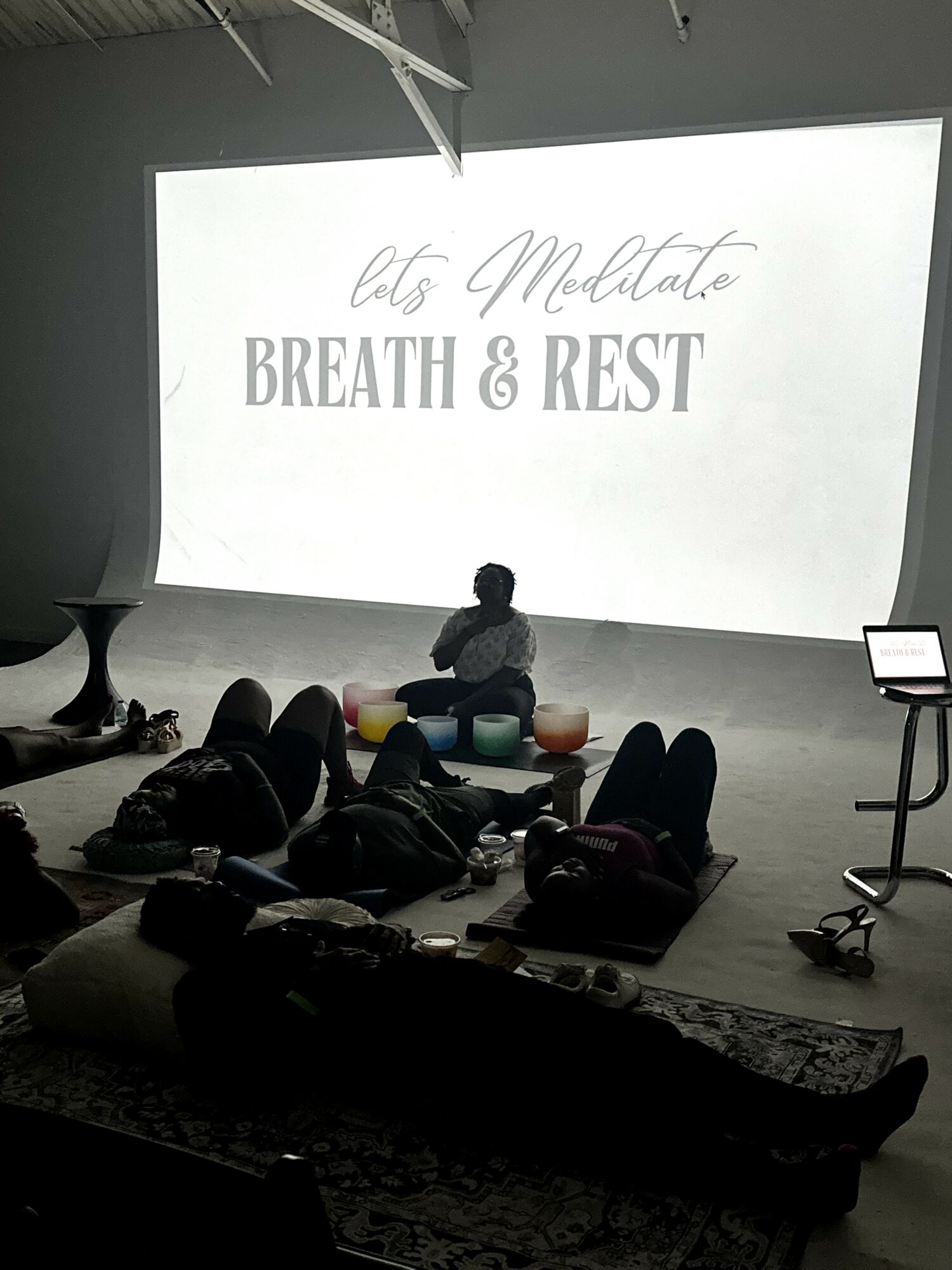 Person sitting cross-legged on floor with meditation cushions, audience lying on mats, large screen displays 'Let's Meditate BREATH & REST'.