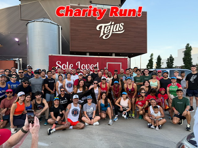 Large group of people gathered outdoors in front of signs, some wearing sunglasses and casual clothing, participating in a charity run.