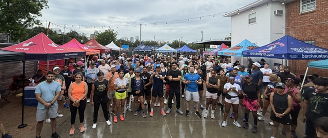 Large group of people gathered outdoors, some standing, some walking, with tents and buildings in the background.