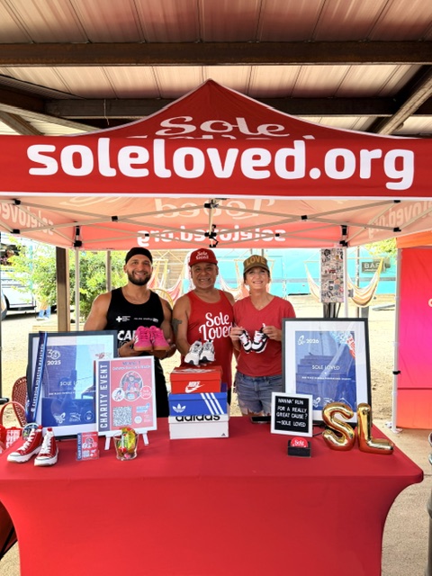 Three people stand behind a table under a red canopy with 'soleloving.org' logo, displaying promotional materials and merchandise.