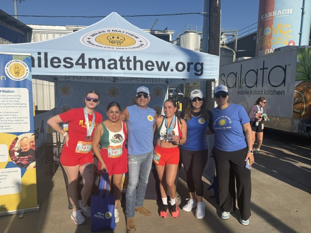 Group of six people standing outdoors in front of a tent and banners, smiling, wearing athletic clothing and sunglasses.