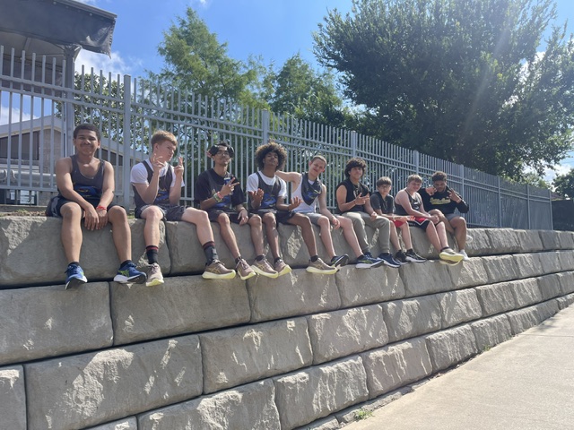 Seven children sit on a stone ledge outdoors, with a fence, trees, and a blue sky in the background.