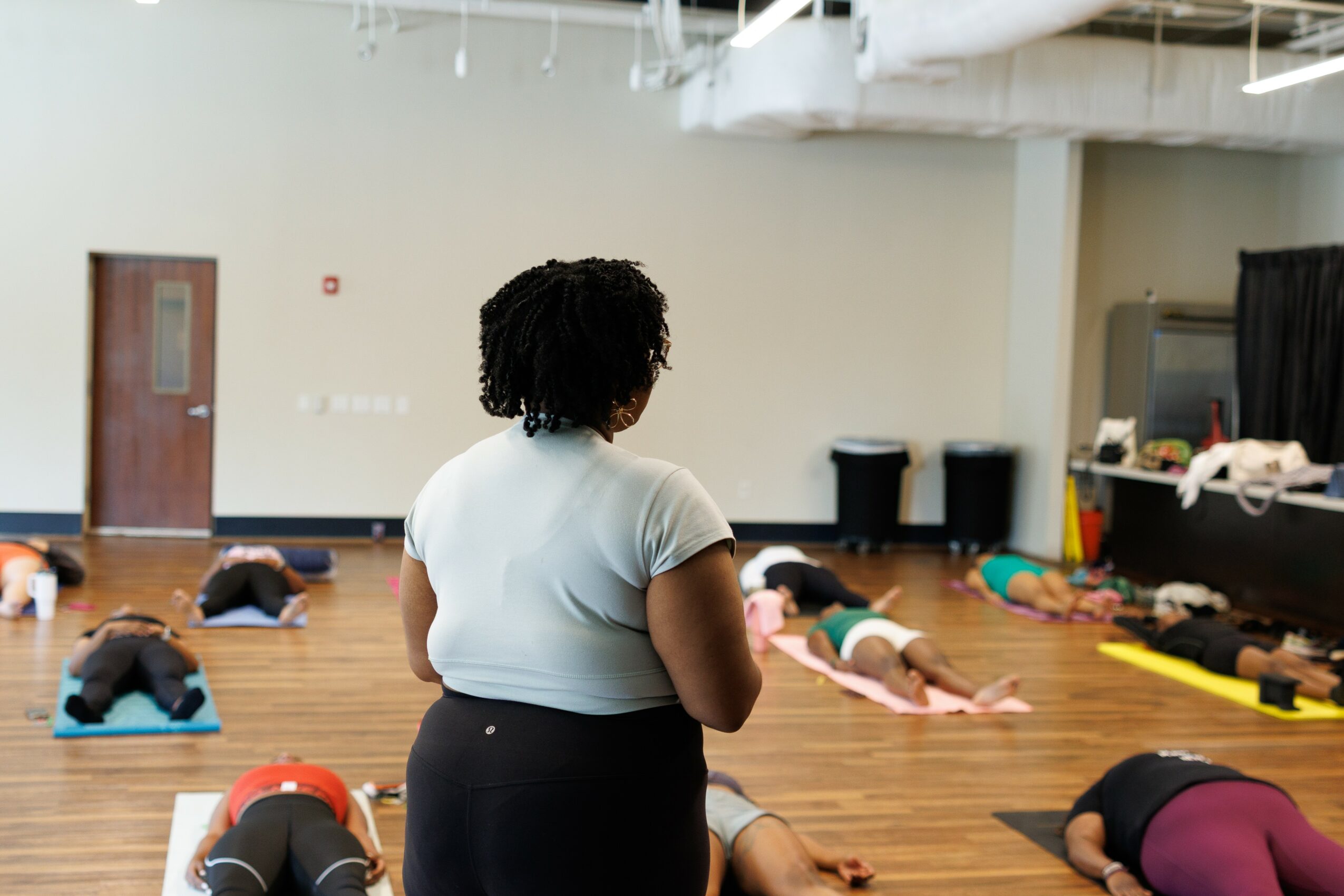 A woman with curly hair stands in a room with people lying on the floor, some on mats, in a fitness or dance class.