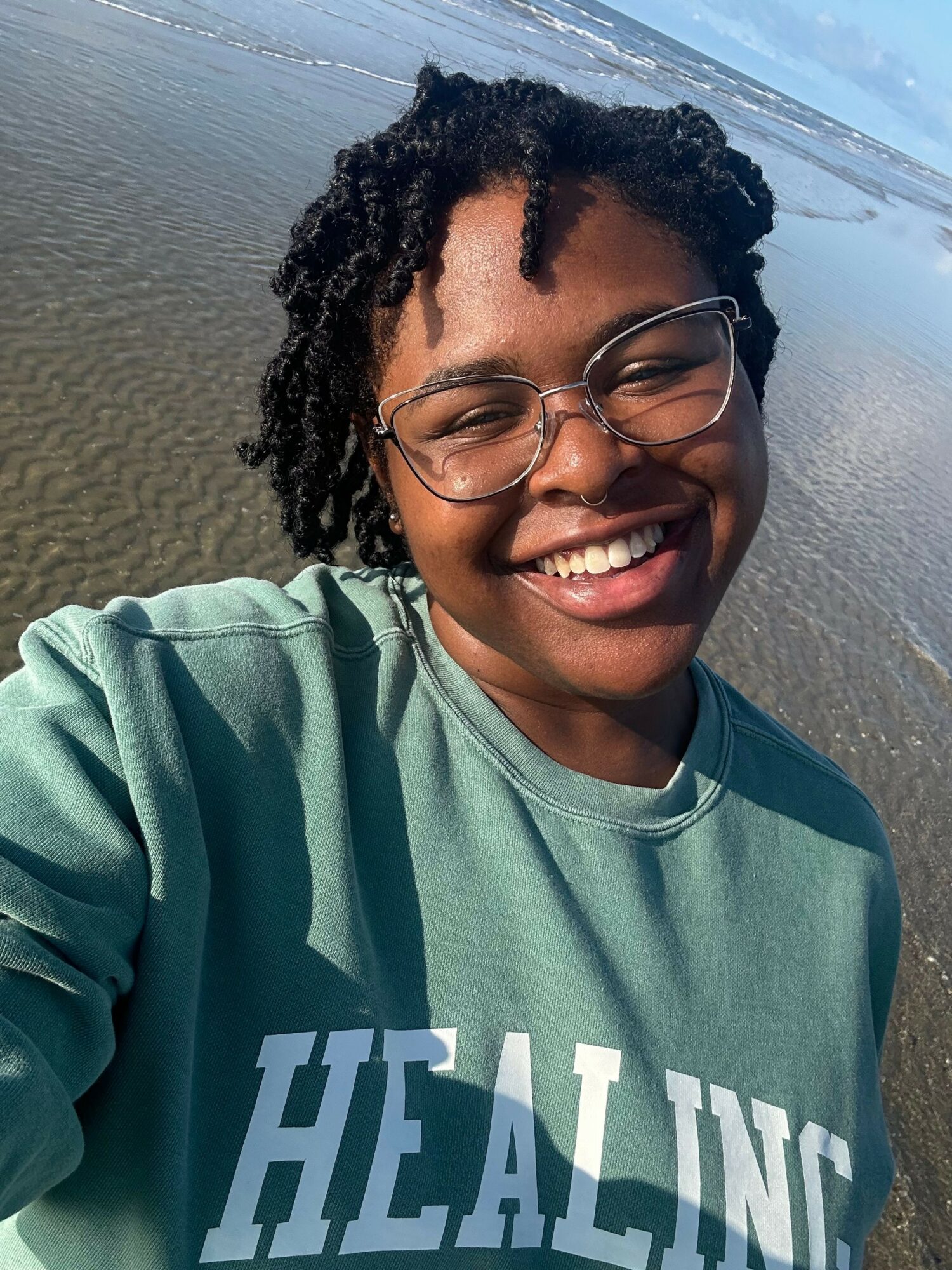 Person with glasses smiling, wearing a green shirt, at the beach with sand and water in the background.