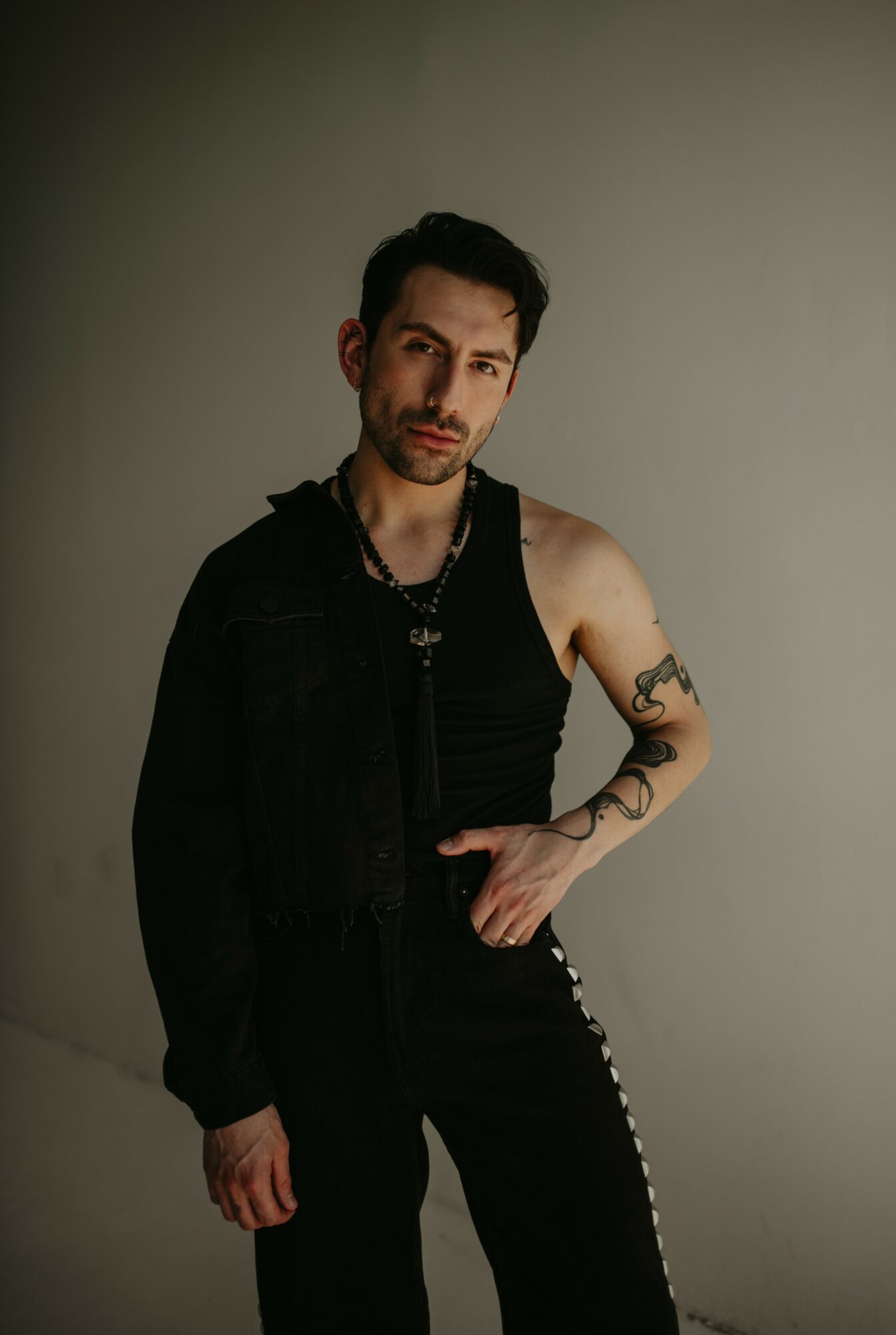 Young man with dark hair and beard, wearing a black sleeveless top and pants, standing against a plain wall.