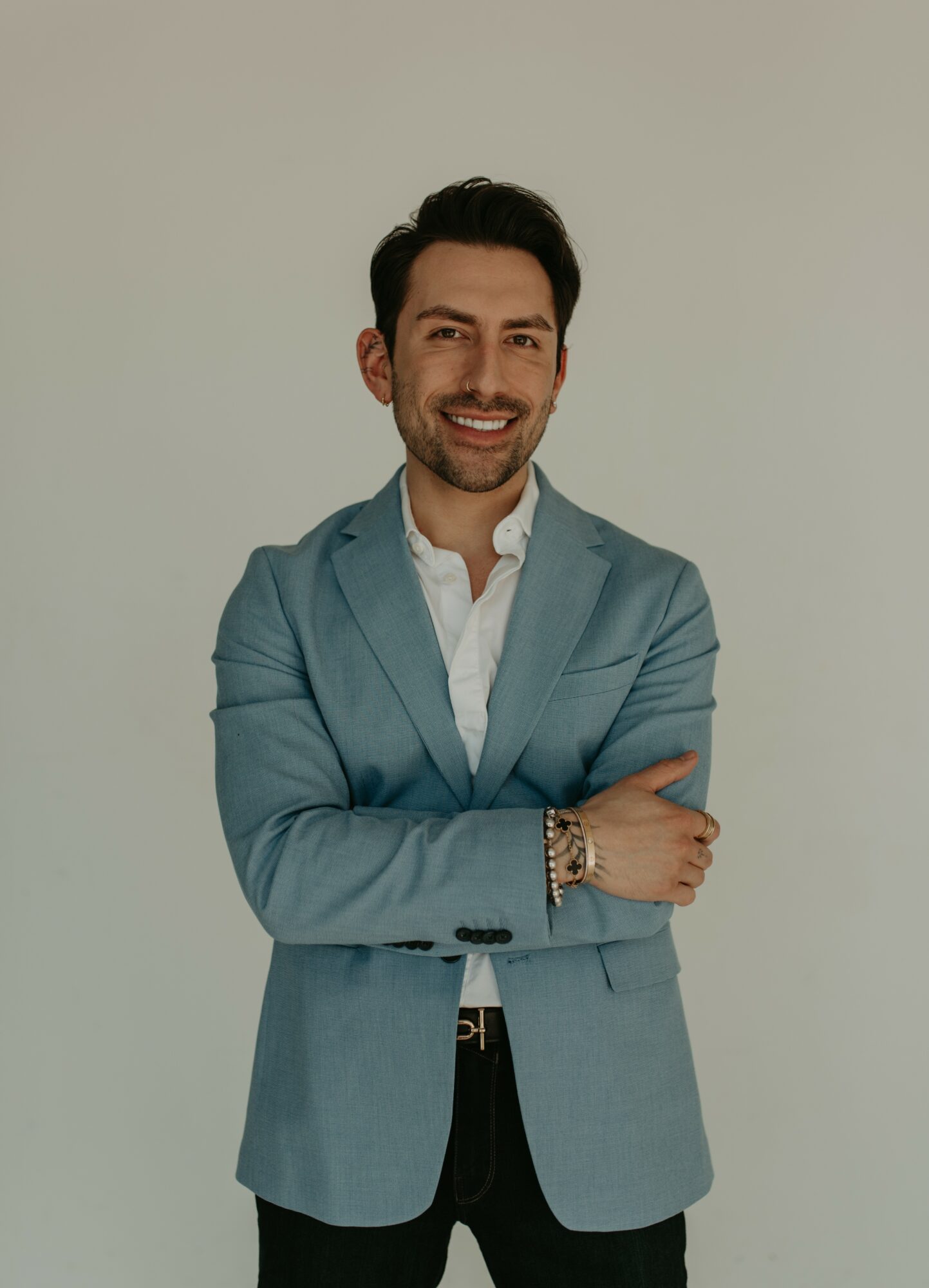 Man in a light blue blazer and white shirt smiling with arms crossed, standing against a plain background.