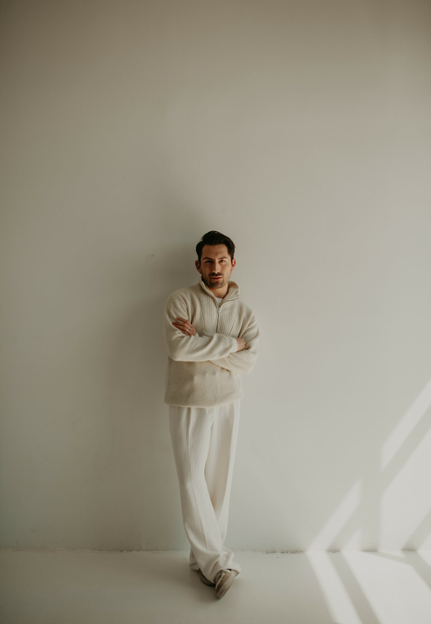 Person standing with arms crossed against a plain wall, with sunlight coming from the right side.