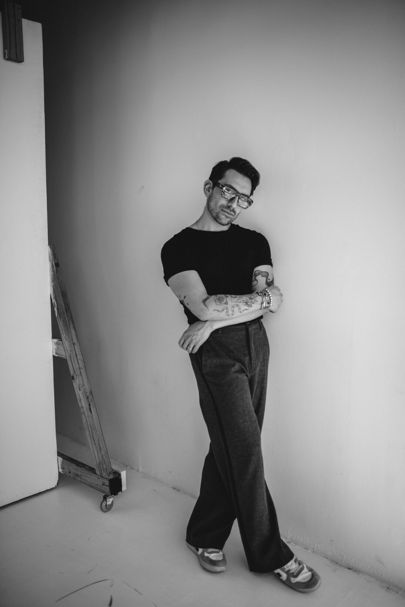 Person with glasses and tattoos leaning against a wall, arms crossed, in black and white. 20 words.