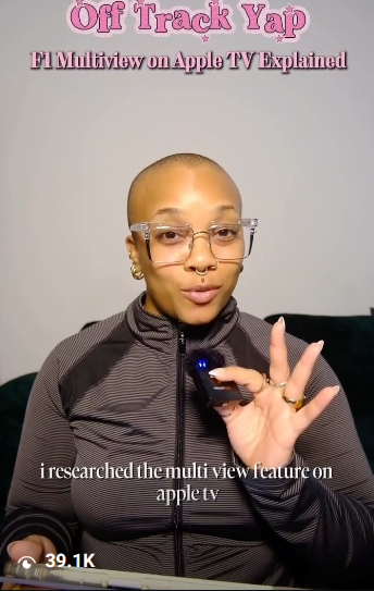 Person with glasses and a shaved head holding a remote, wearing a dark jacket, in front of a light background.