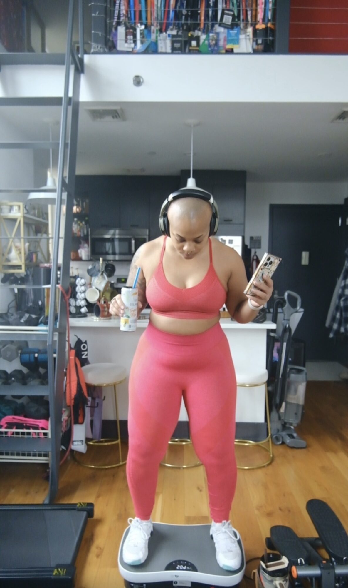 Woman in pink workout clothes listening to headphones and looking at her phone in a modern home gym.