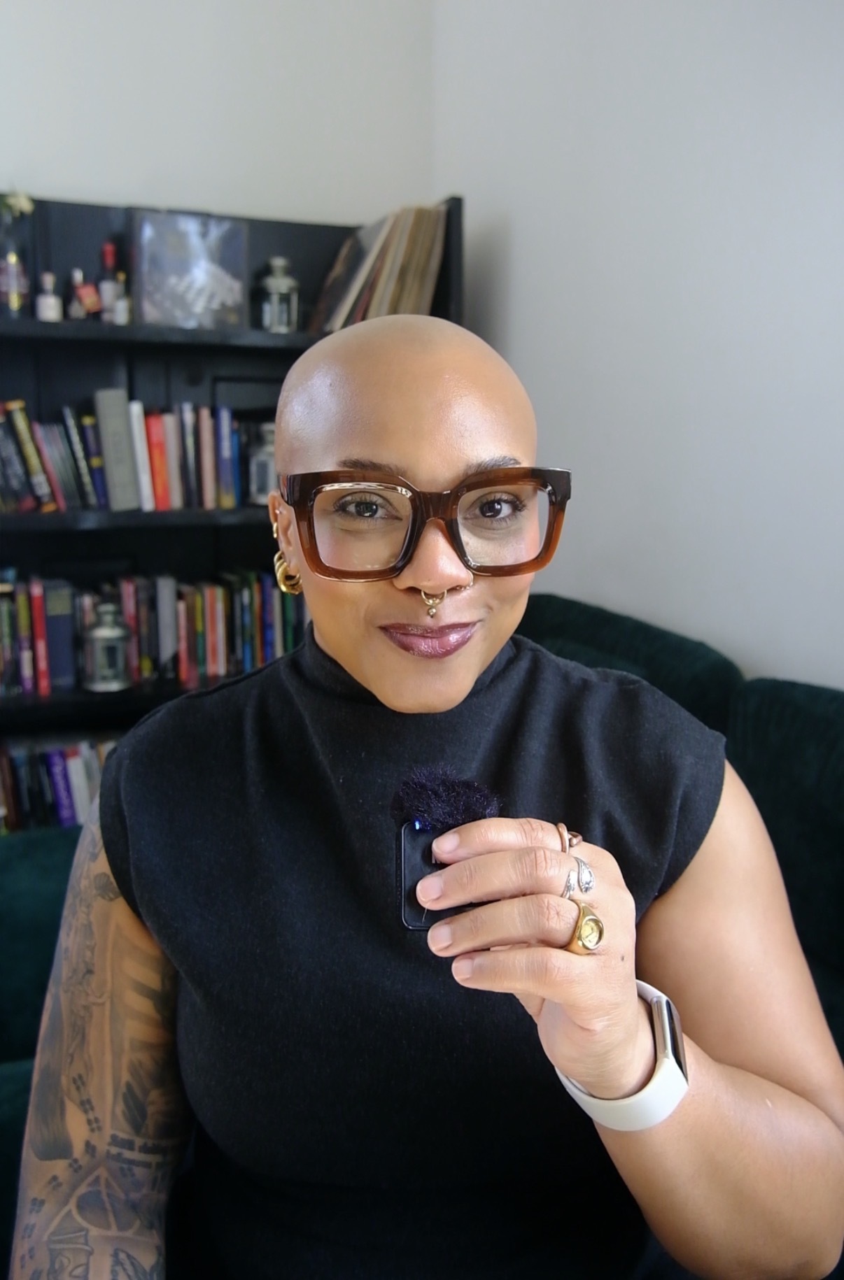 Bald woman with glasses and a black sleeveless top holding a phone, standing in front of a bookshelf.