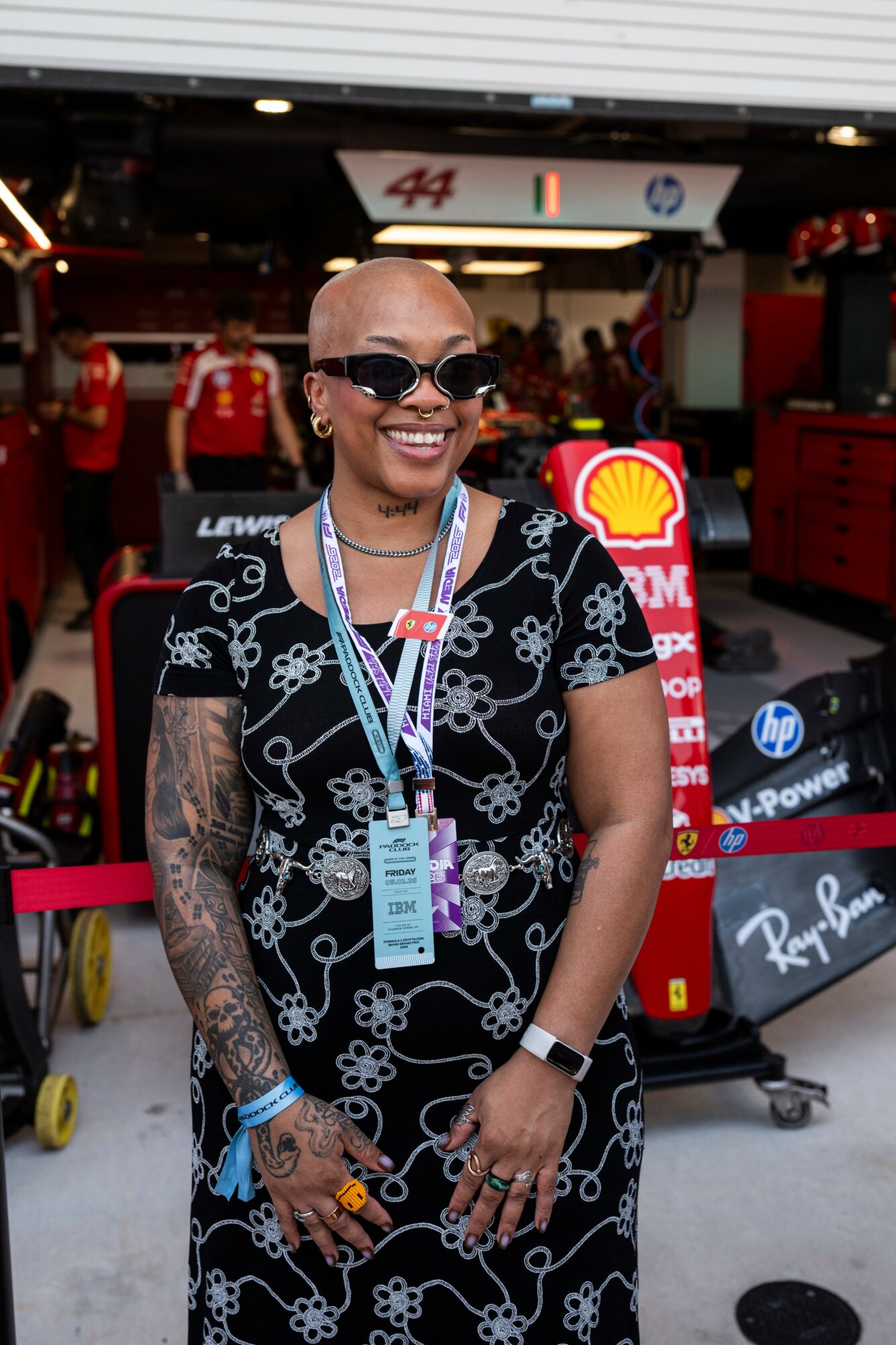 Smiling woman with tattoos and sunglasses standing in a racing garage, wearing a black dress with white floral patterns, with racing team members and equipment in the background.