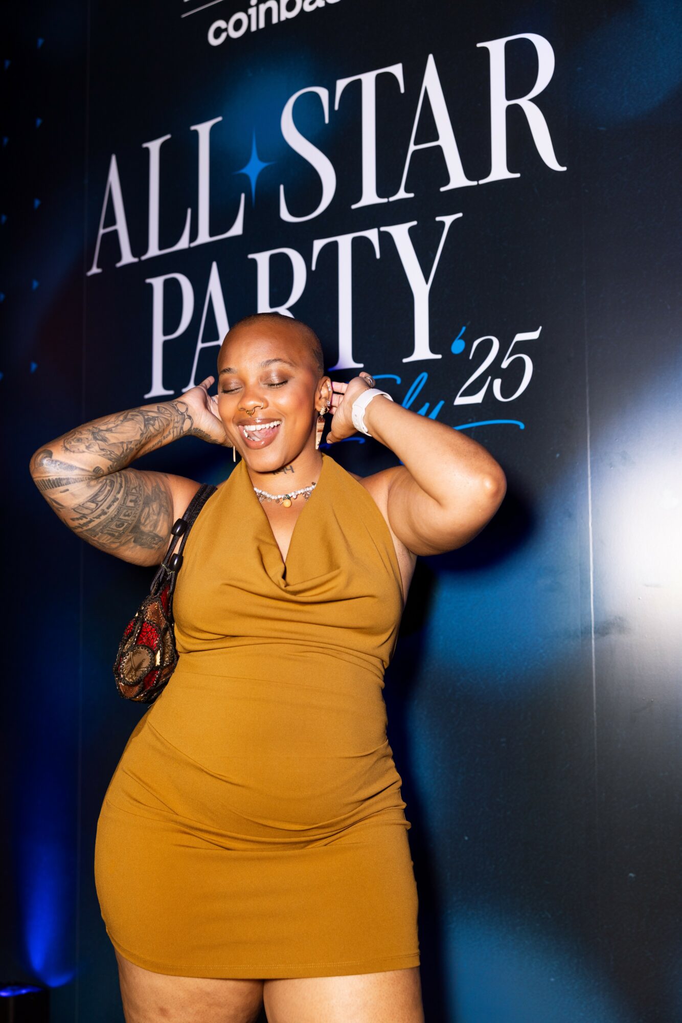 Smiling woman with tattoos and jewelry poses at event, standing in front of a sign that reads 'ALL STAR PARTY'.