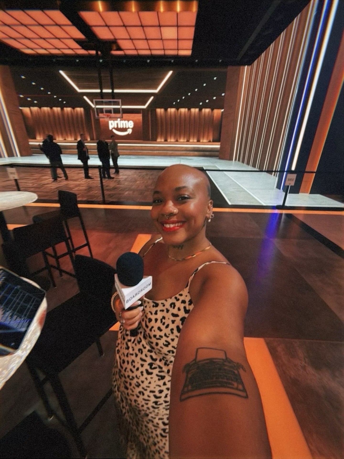 Woman smiling, holding microphone, in a large, modern event space with a stage and people in the background.
