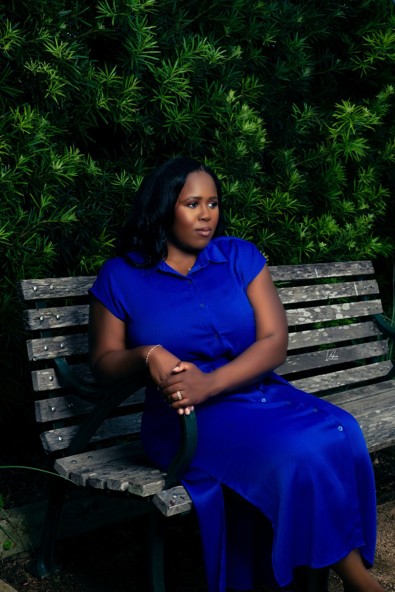 Woman in blue dress sitting on a wooden bench with green foliage background.