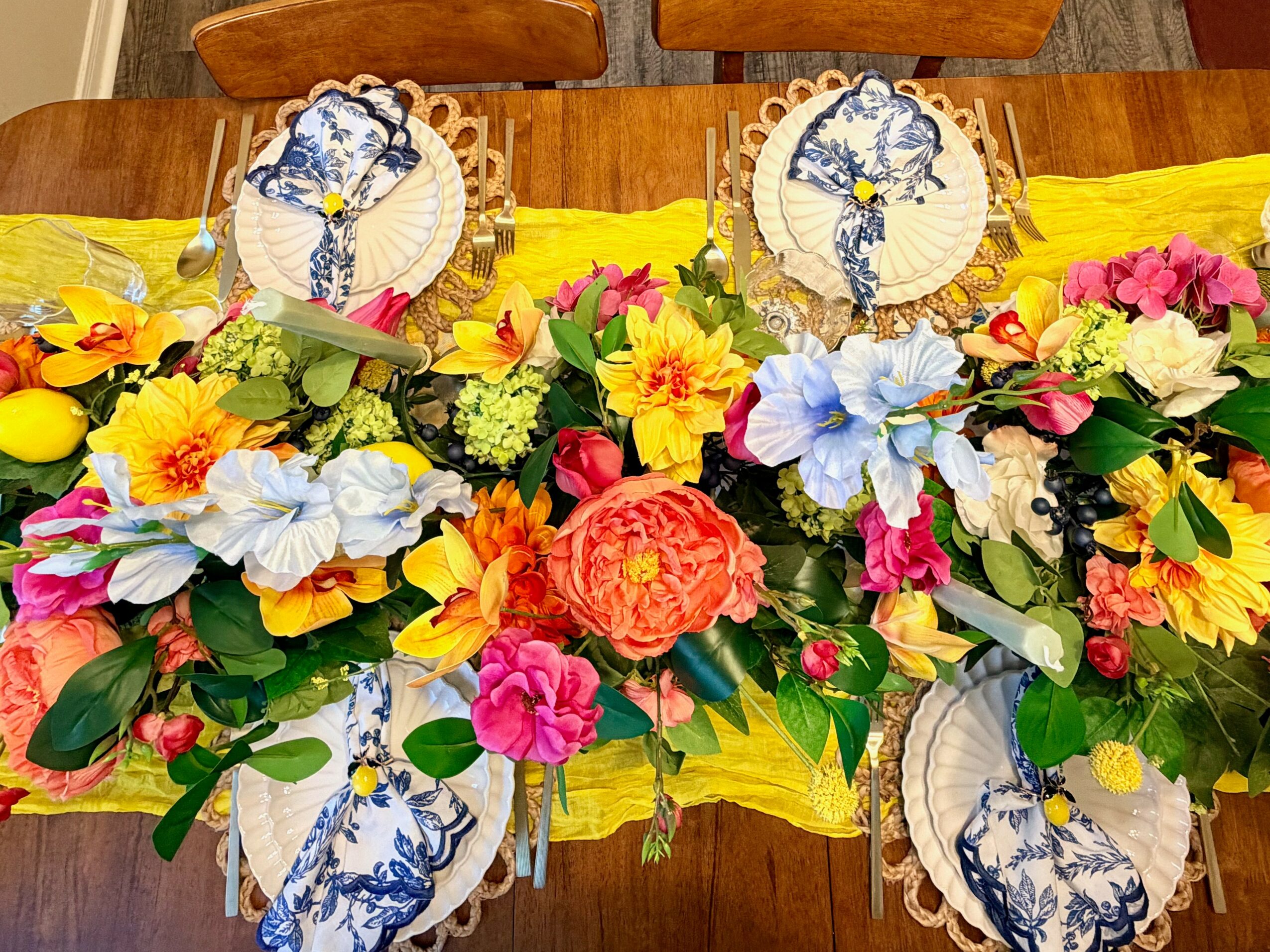 Four floral arrangements with yellow, pink, blue, and white flowers on a wooden table with decorative plates and yellow table runner.