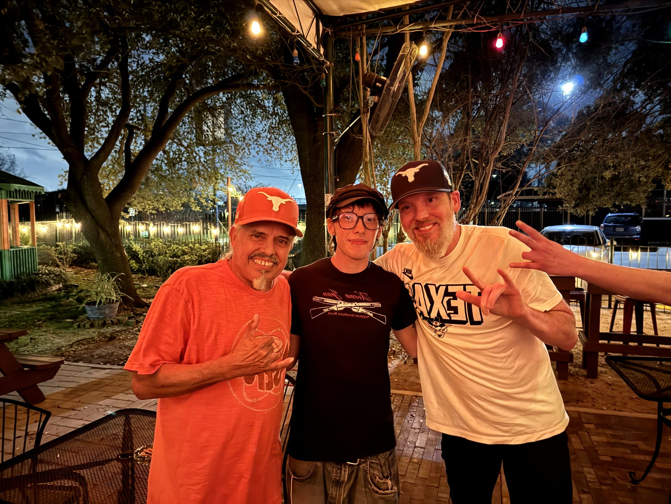 Three people standing outdoors at night, smiling, with trees and string lights in the background.
