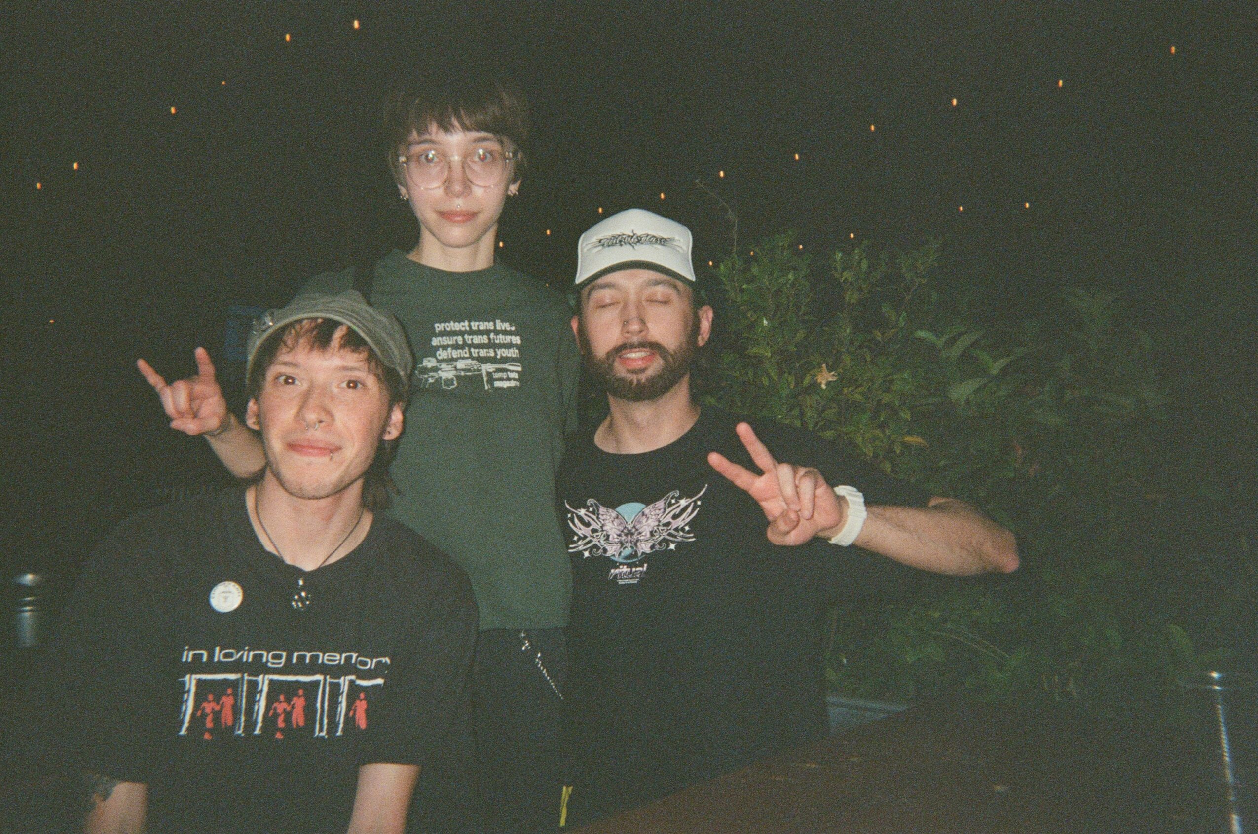Three people posing outdoors at night, two making peace signs, one standing behind them, all smiling.