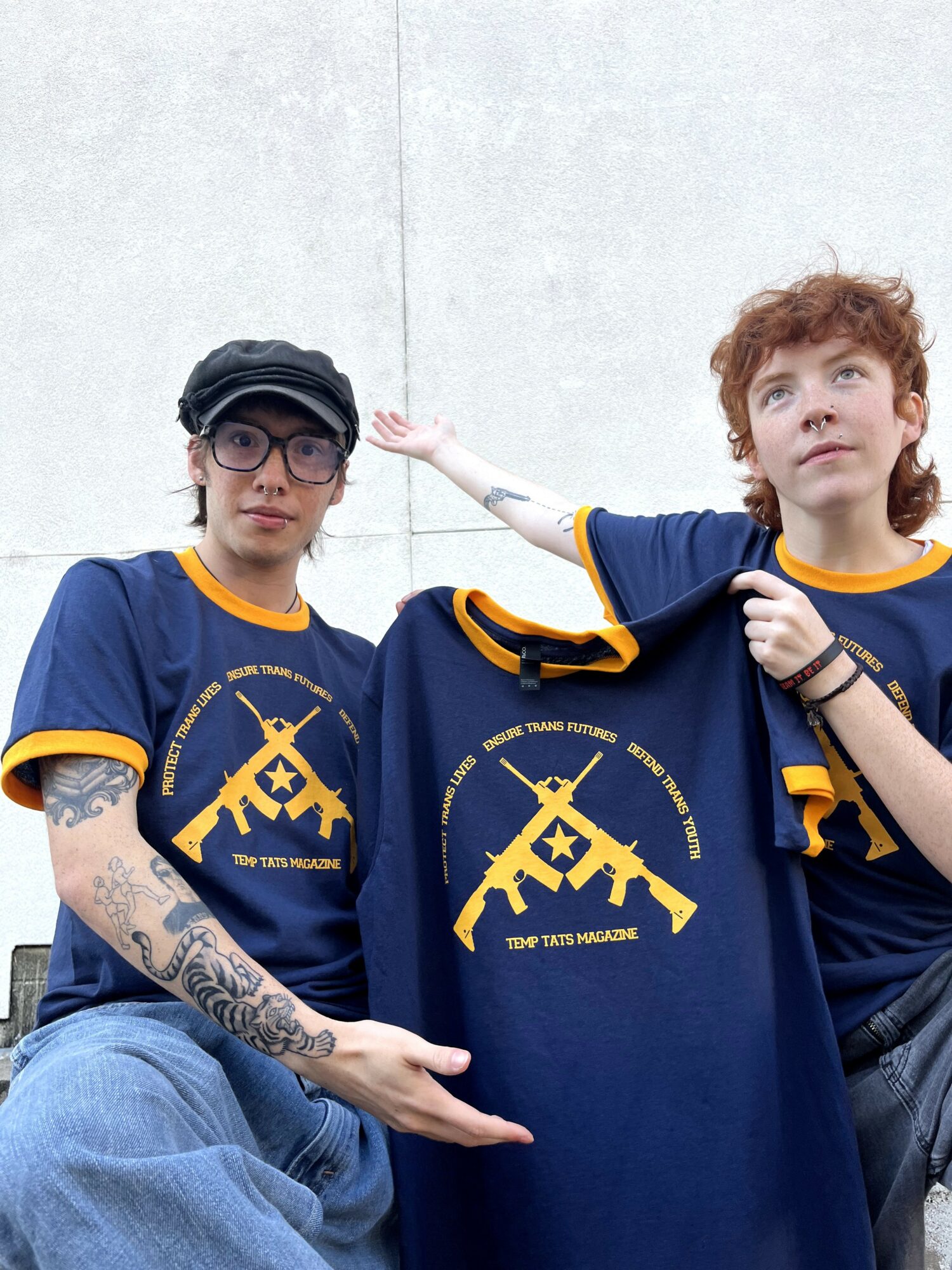Two young people in blue and yellow sports jerseys holding a matching jersey, one with tattoos and glasses, the other with curly hair, against a plain background.