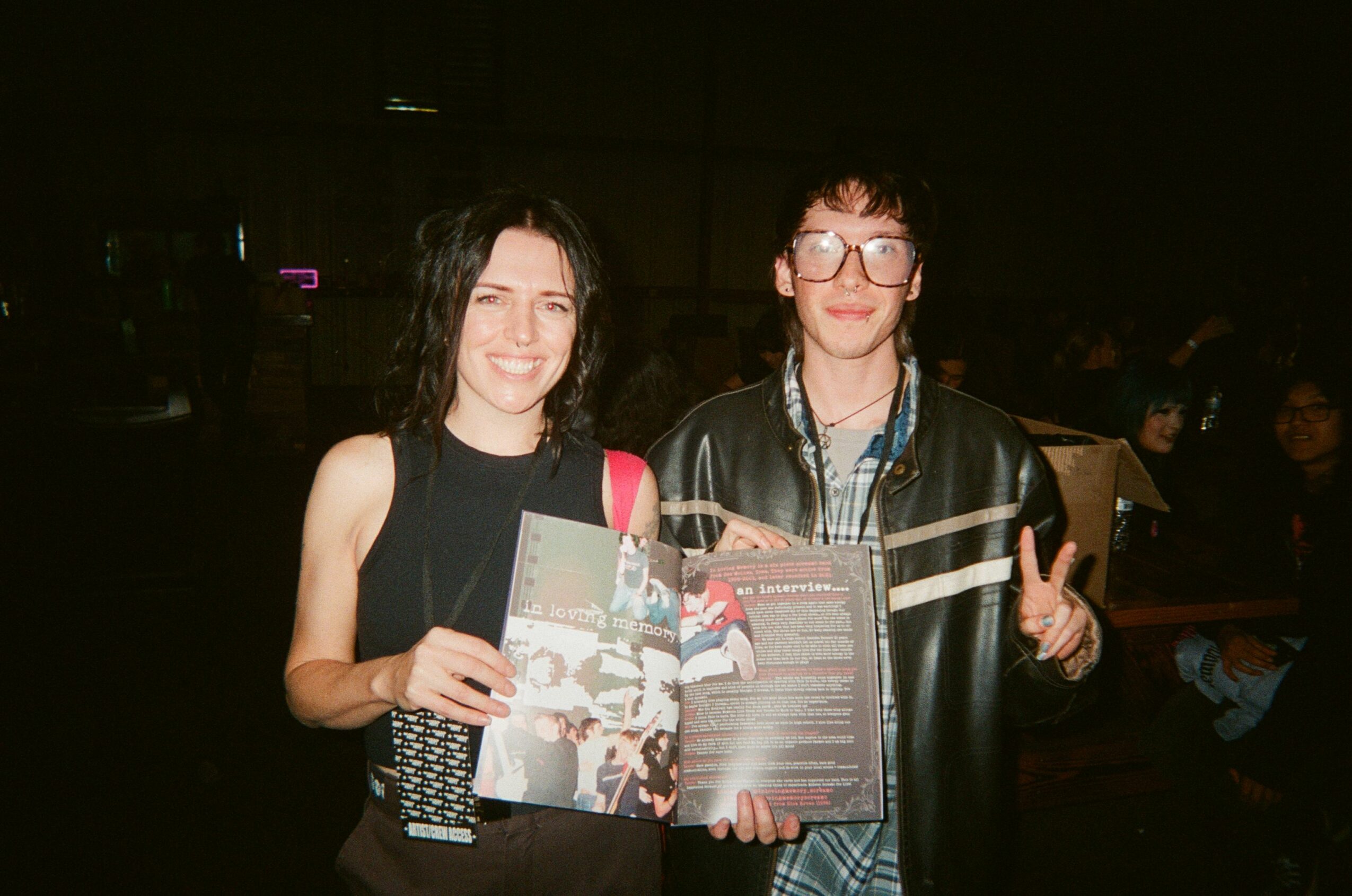 Two women sitting at a table, one holding a magazine, smiling, in a dimly lit environment.