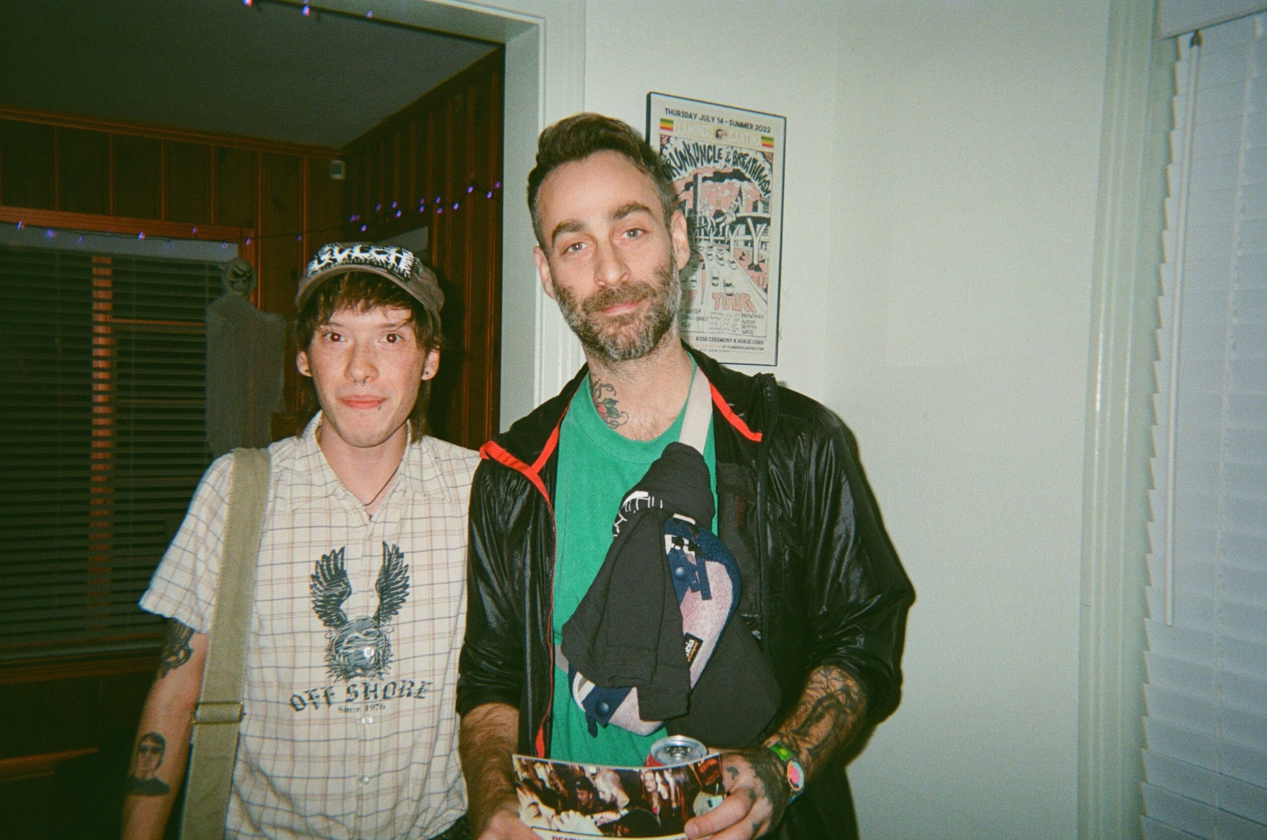 Two people standing indoors, one with a cap and casual clothing, the other with a beard and leather jacket, smiling.
