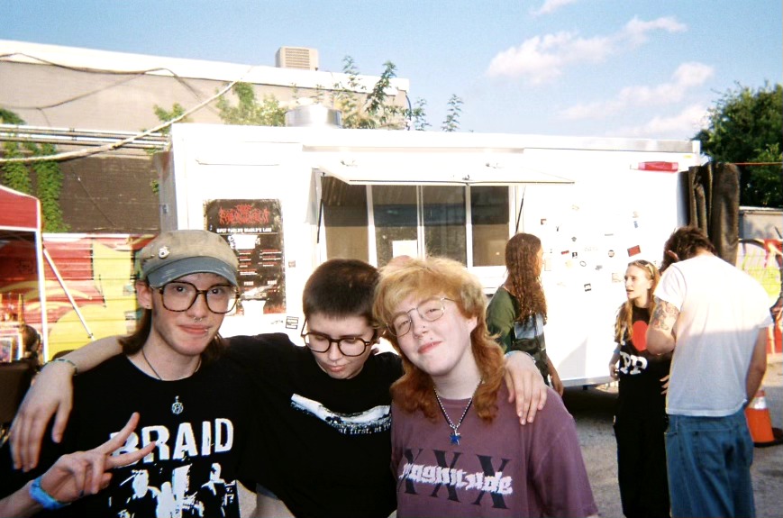 Three people with arms around each other in front of a food truck, smiling outdoors on a sunny day.