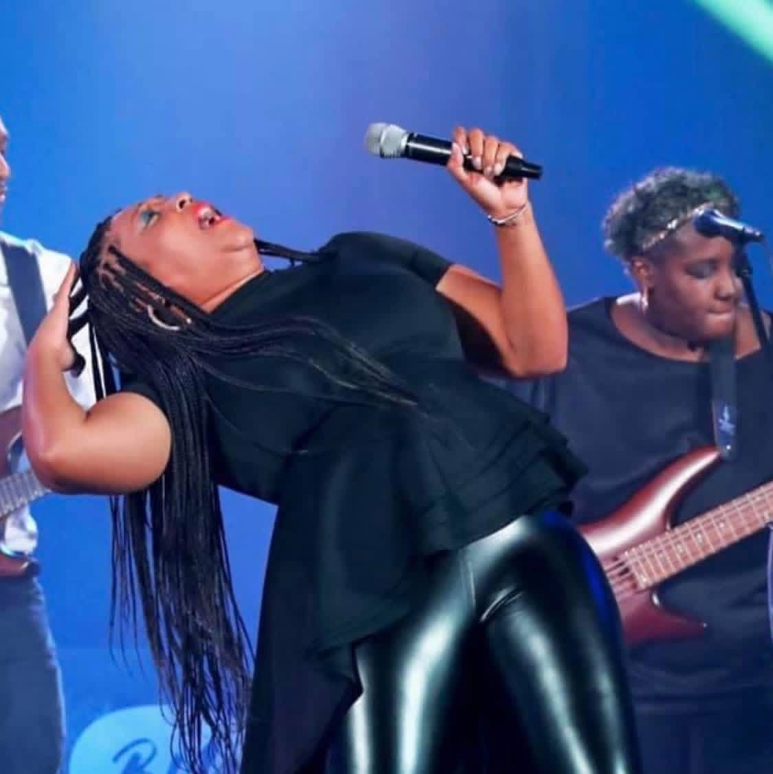 Woman singing into a microphone on stage with a guitarist in the background, blue lighting, and a dark outfit.