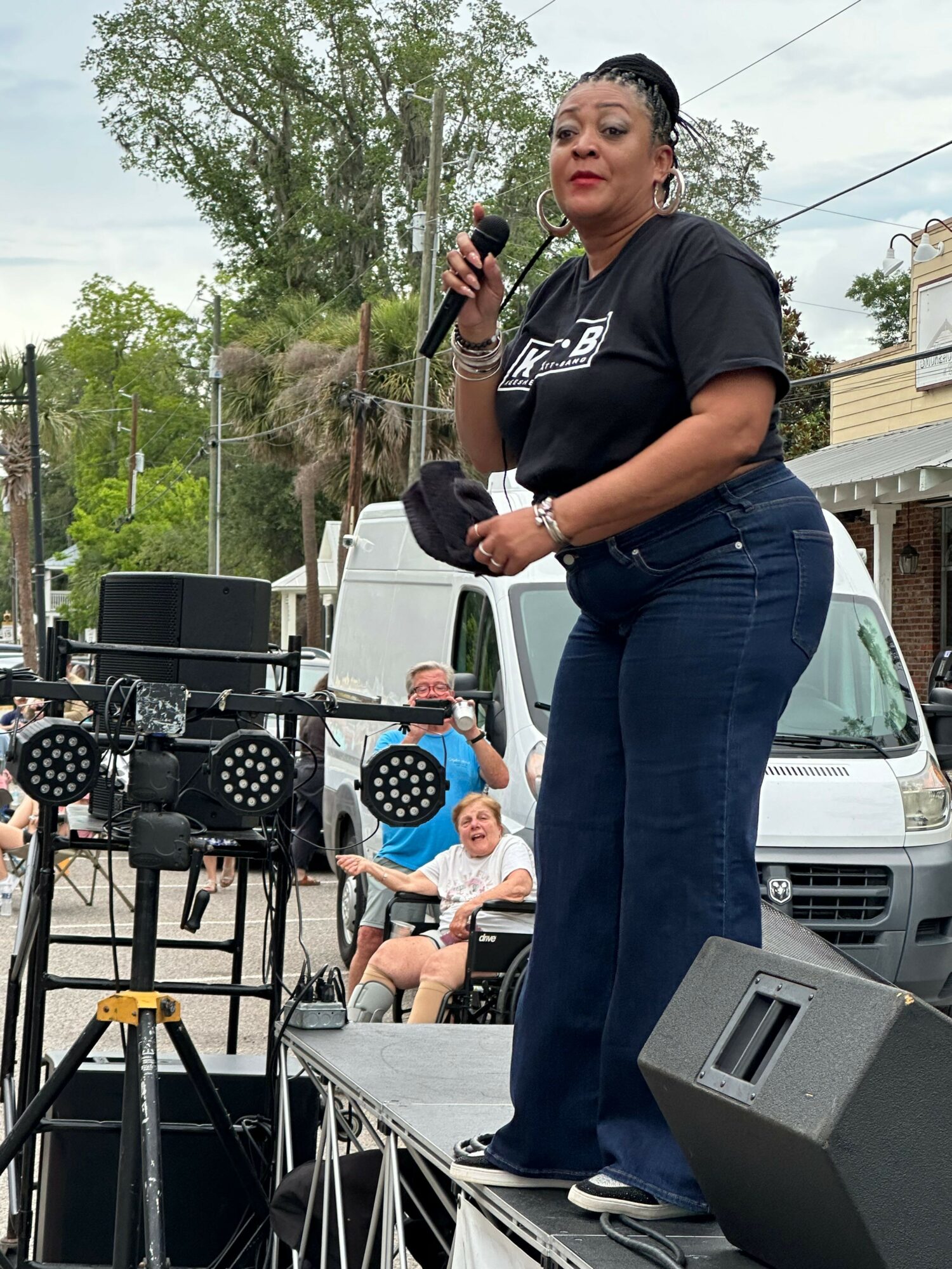 Woman speaking into microphone outdoors with people and trees in background.