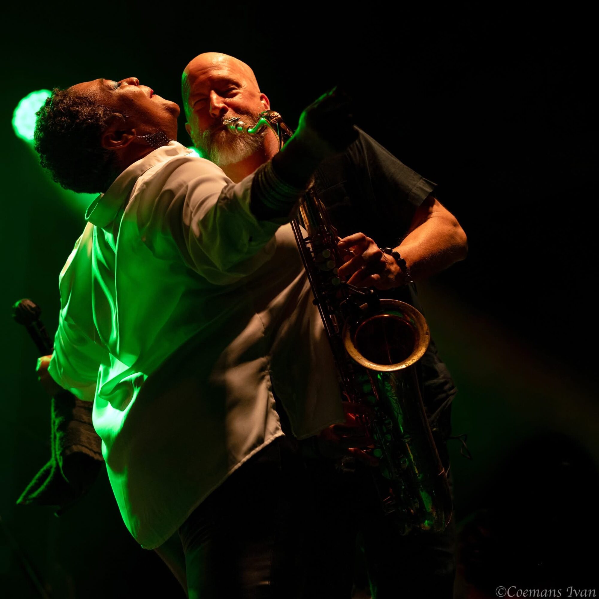 Two musicians perform on stage, one singing and the other playing a saxophone, illuminated by green and yellow stage lights.