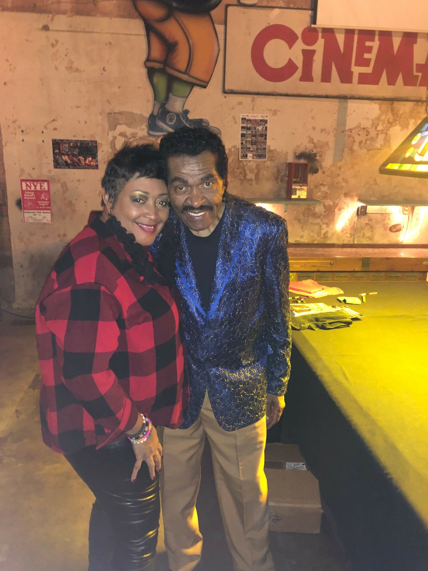 Two women standing close together in a dimly lit indoor setting, smiling at the camera, with a wall and posters behind them.