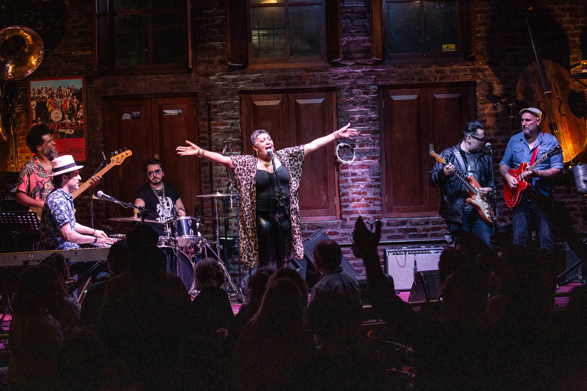 Musicians perform on stage with a singer in the center, audience silhouettes in foreground, brick wall background, warm lighting.