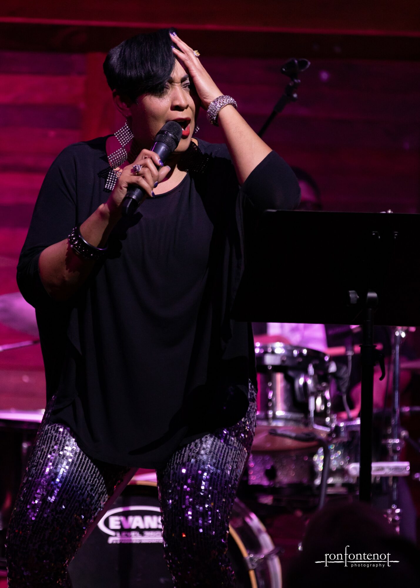 Woman singing into a microphone on stage with musical instruments in the background, wearing a black outfit with sparkly pants.