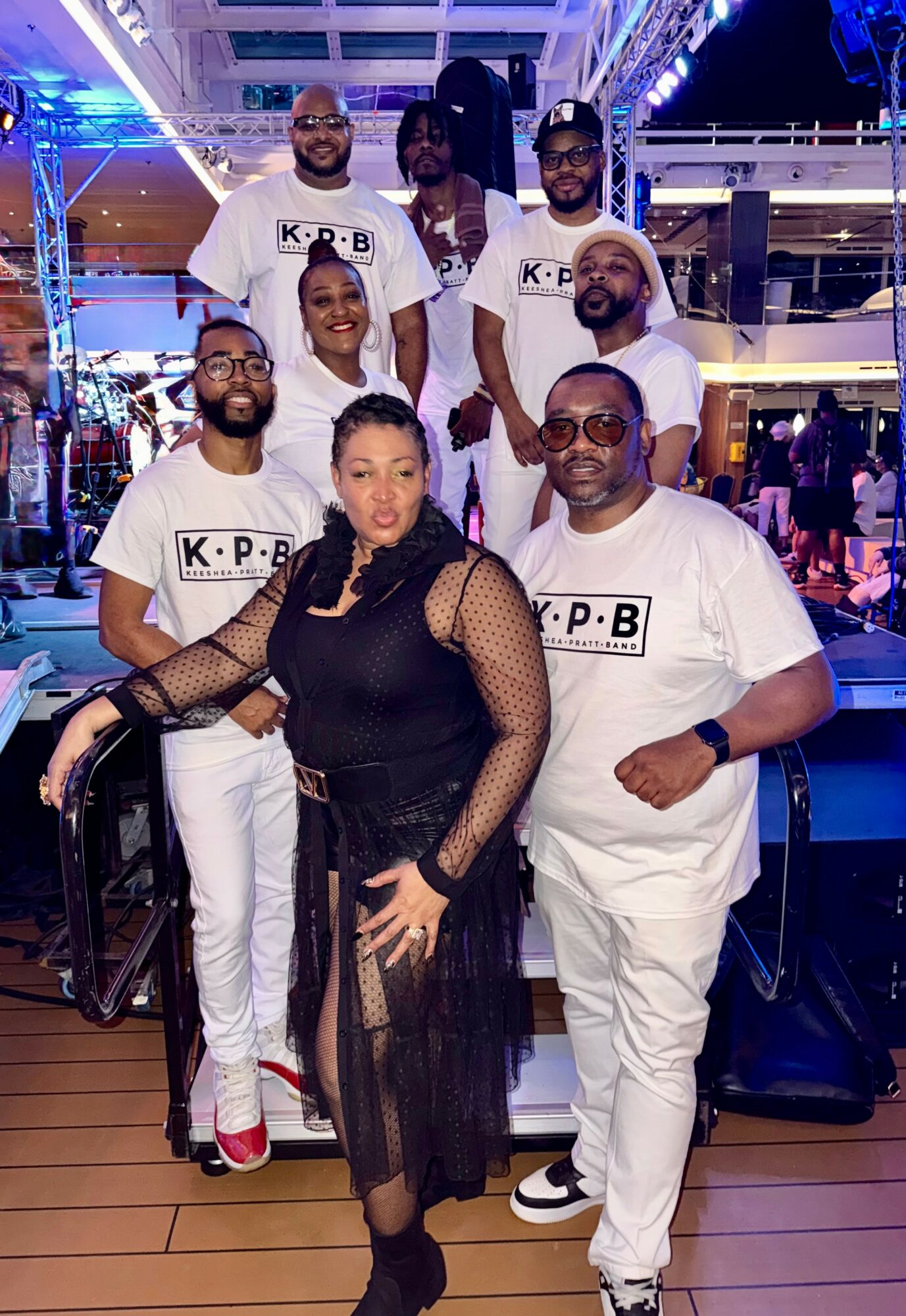 Group of nine people posing indoors with colorful lighting, some wearing matching white T-shirts, one woman in black dress, all smiling.