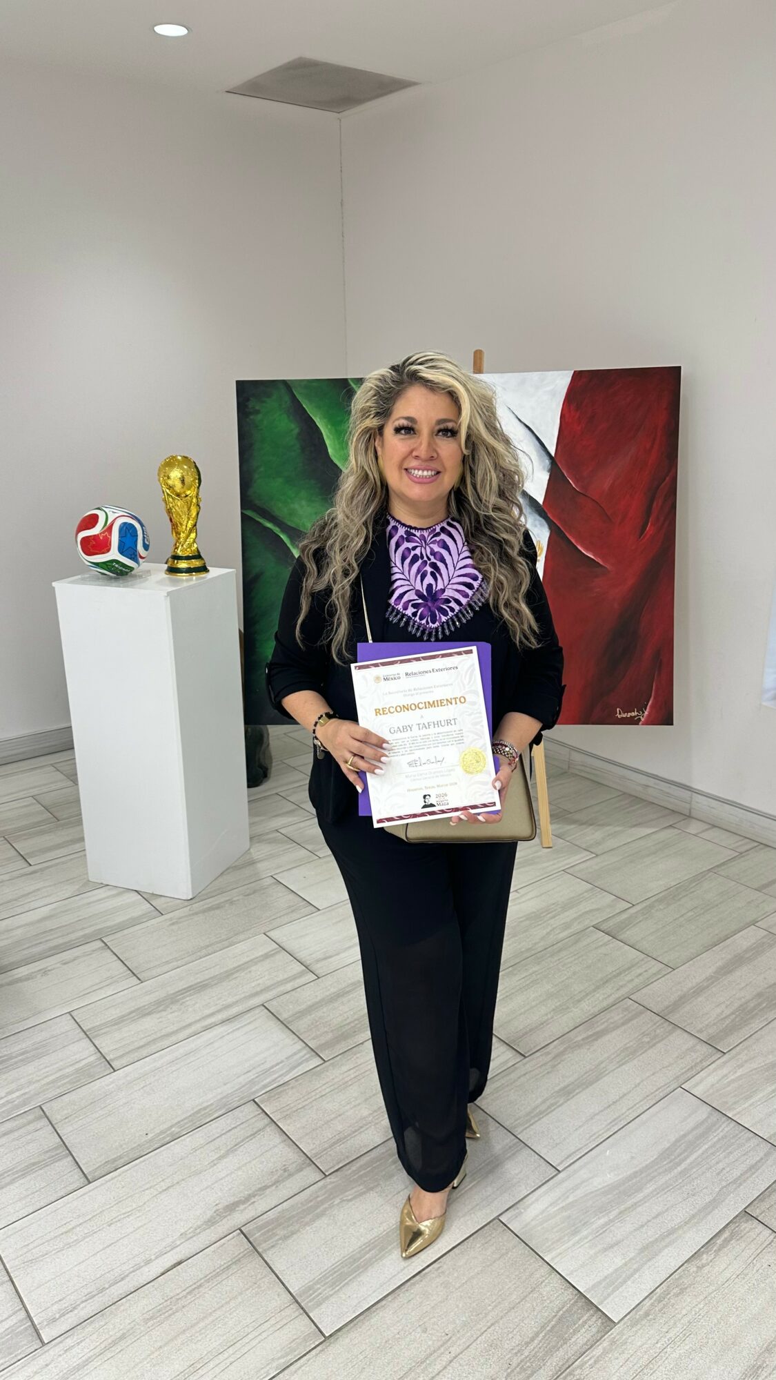Woman holding a certificate, standing in an art gallery with sculptures and paintings, smiling, with curly hair and a purple scarf.