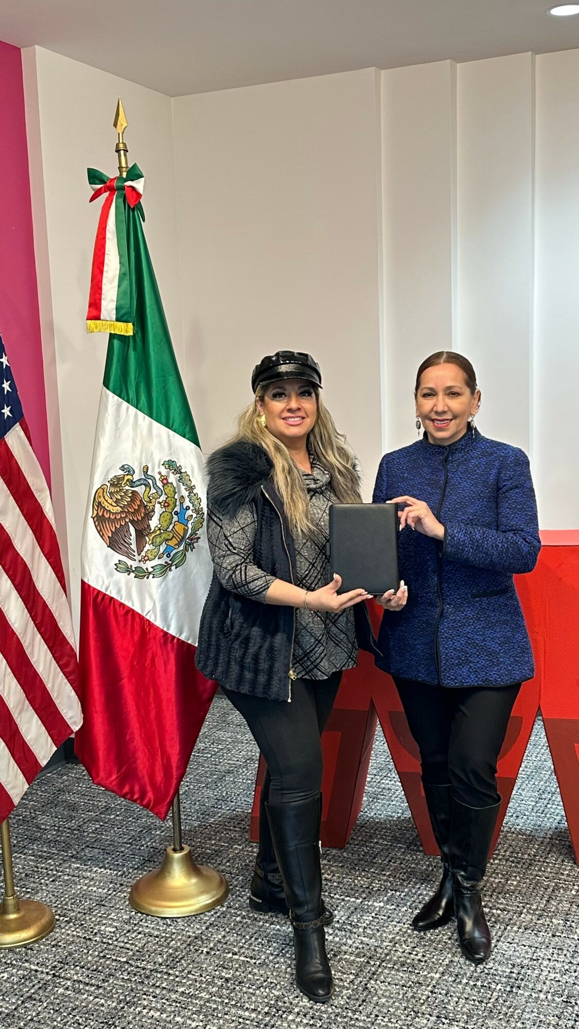 Two women standing indoors, holding a document, with the Mexican flag and American flag behind them.