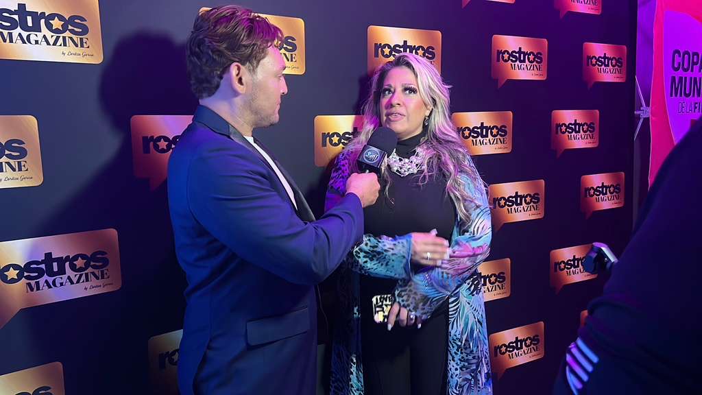 Two people speaking at an event with a backdrop of Rostros Magazine logos, one holding a microphone and a bouquet.