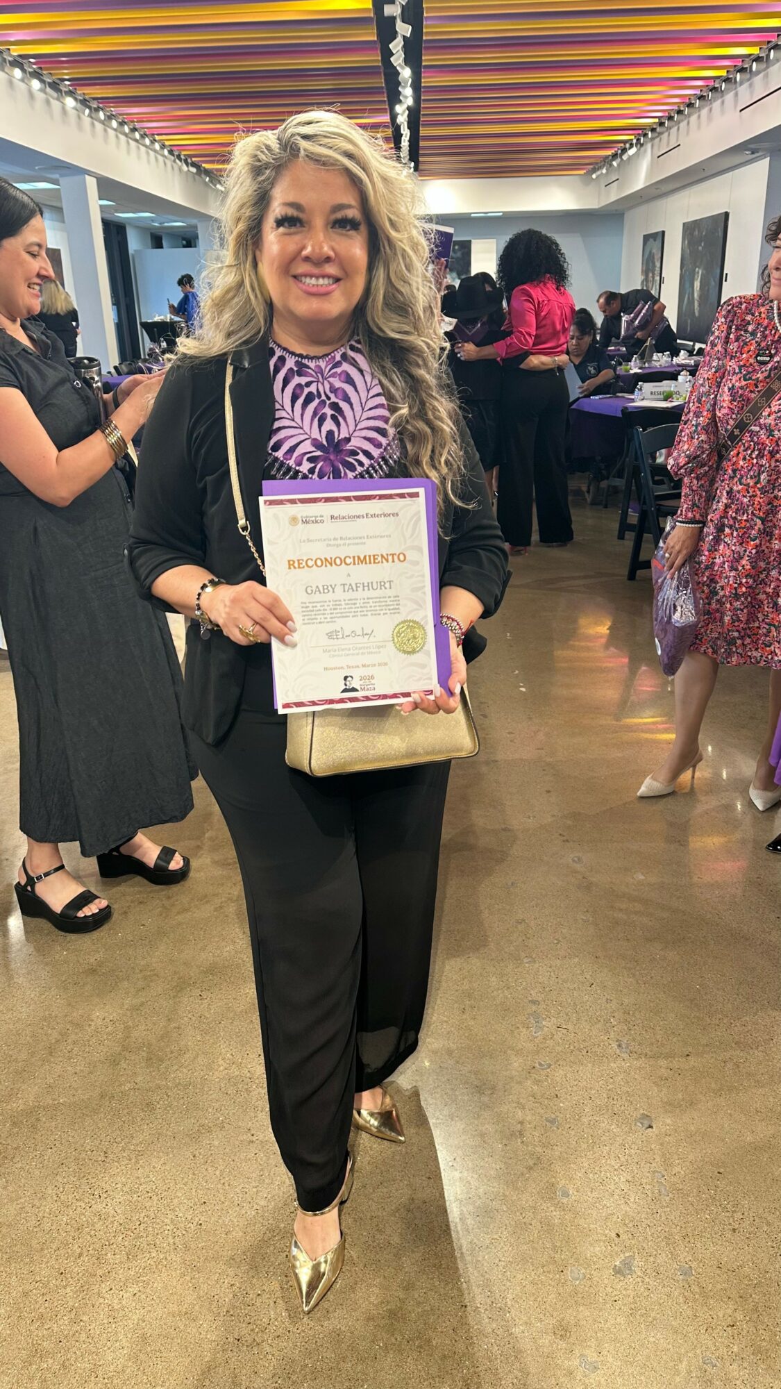Woman with curly blonde hair holding a certificate, smiling, in a colorful indoor setting with other people in the background.